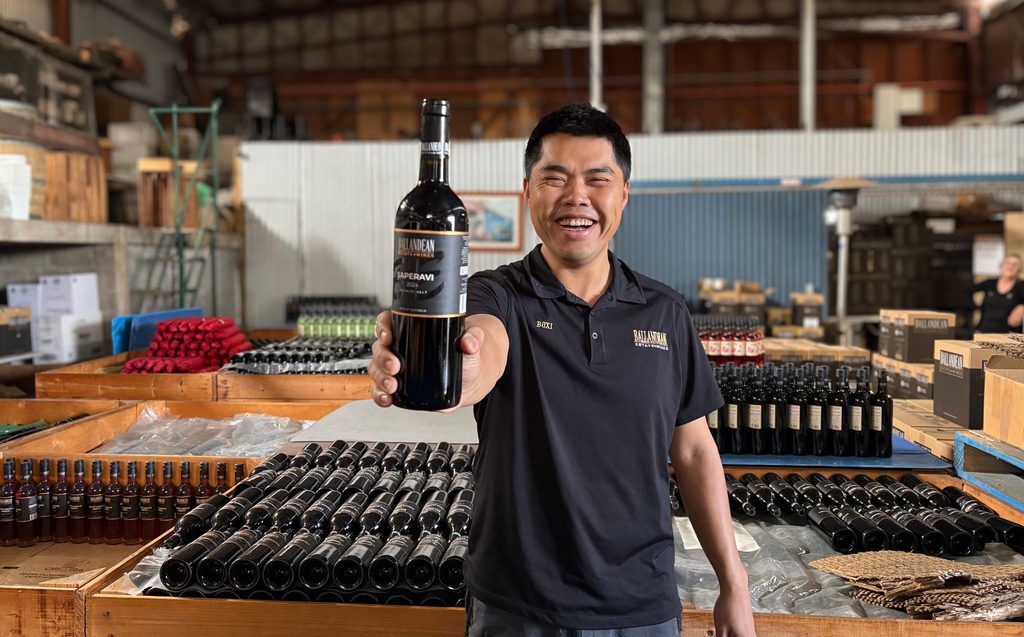 Young male winemaker inside production facility holds award winning bottle of Saperavi in front of him with big smile